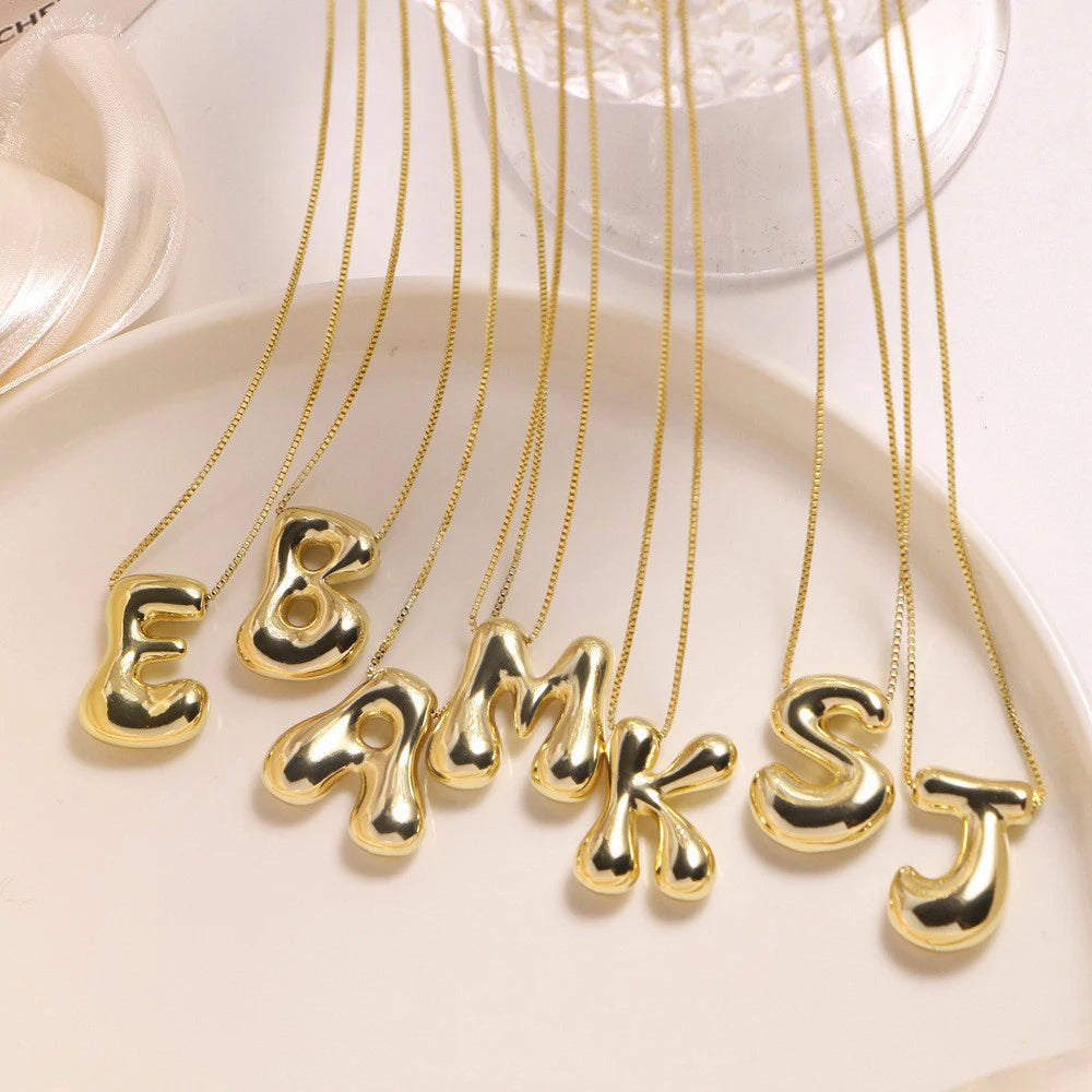 Bold Bubble Initial Pendant Necklace Gold multiple letter variants hanging on beige surface