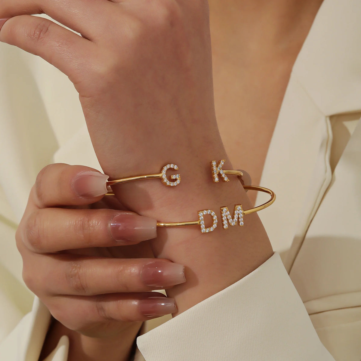 Two gold double initial cuff bracelets with G, K and D, M worn on the wrist