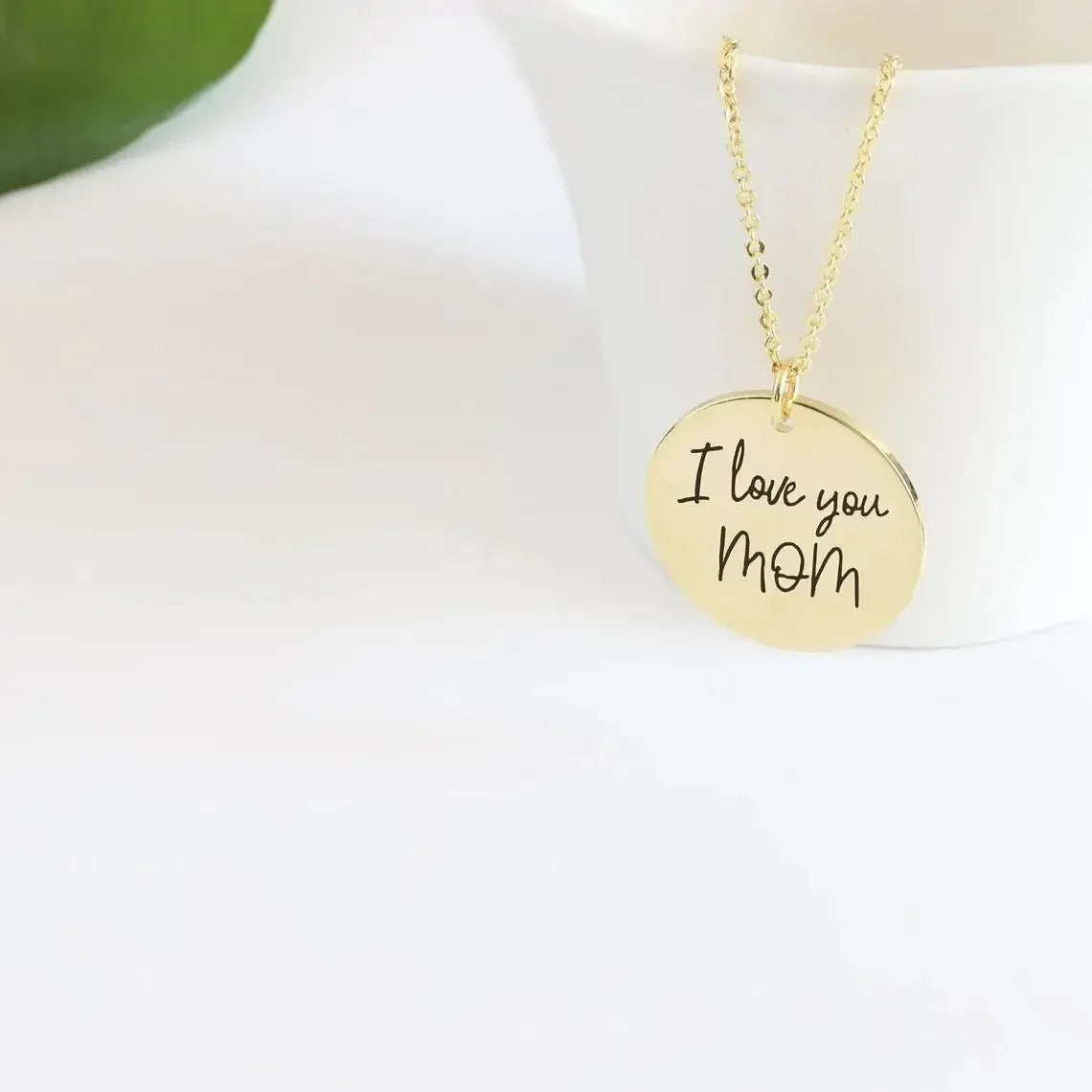 Gold handwriting disc necklace engraved I love you mom hanging from a white cup